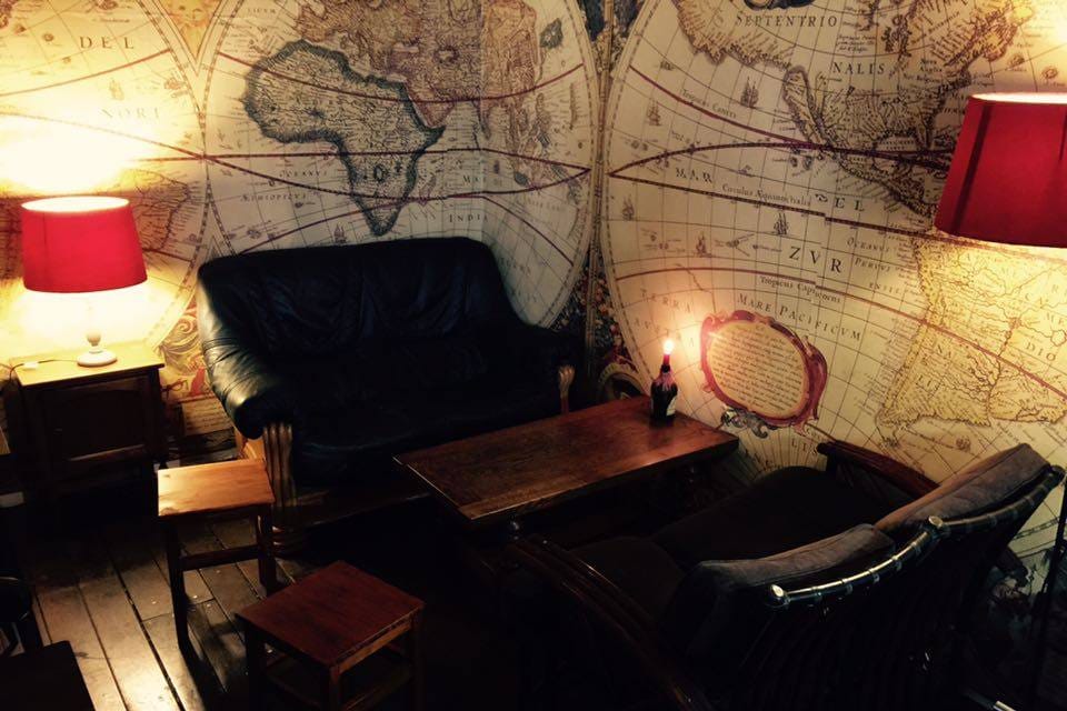 Een hoek van de bovenverdieping met een leren zetel, een rode lamp en houten tafels tegen een muur vol oude kaarten.