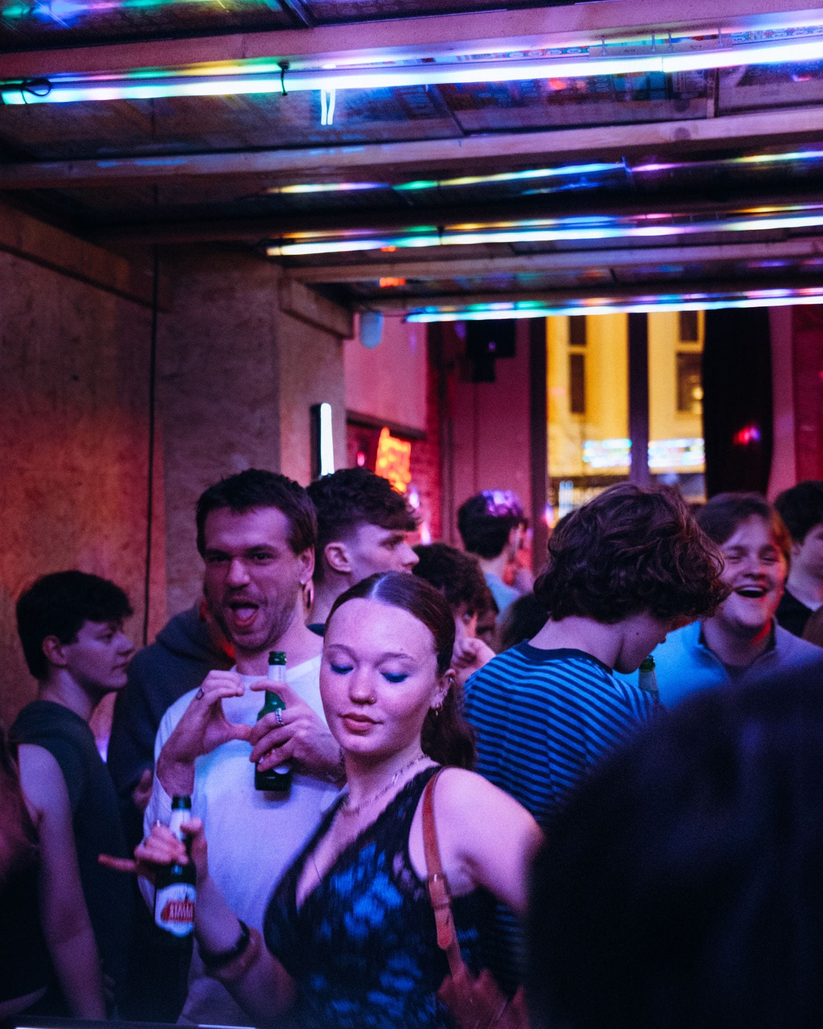 People dancing with bottles in hand under pink and blue neon tubes.