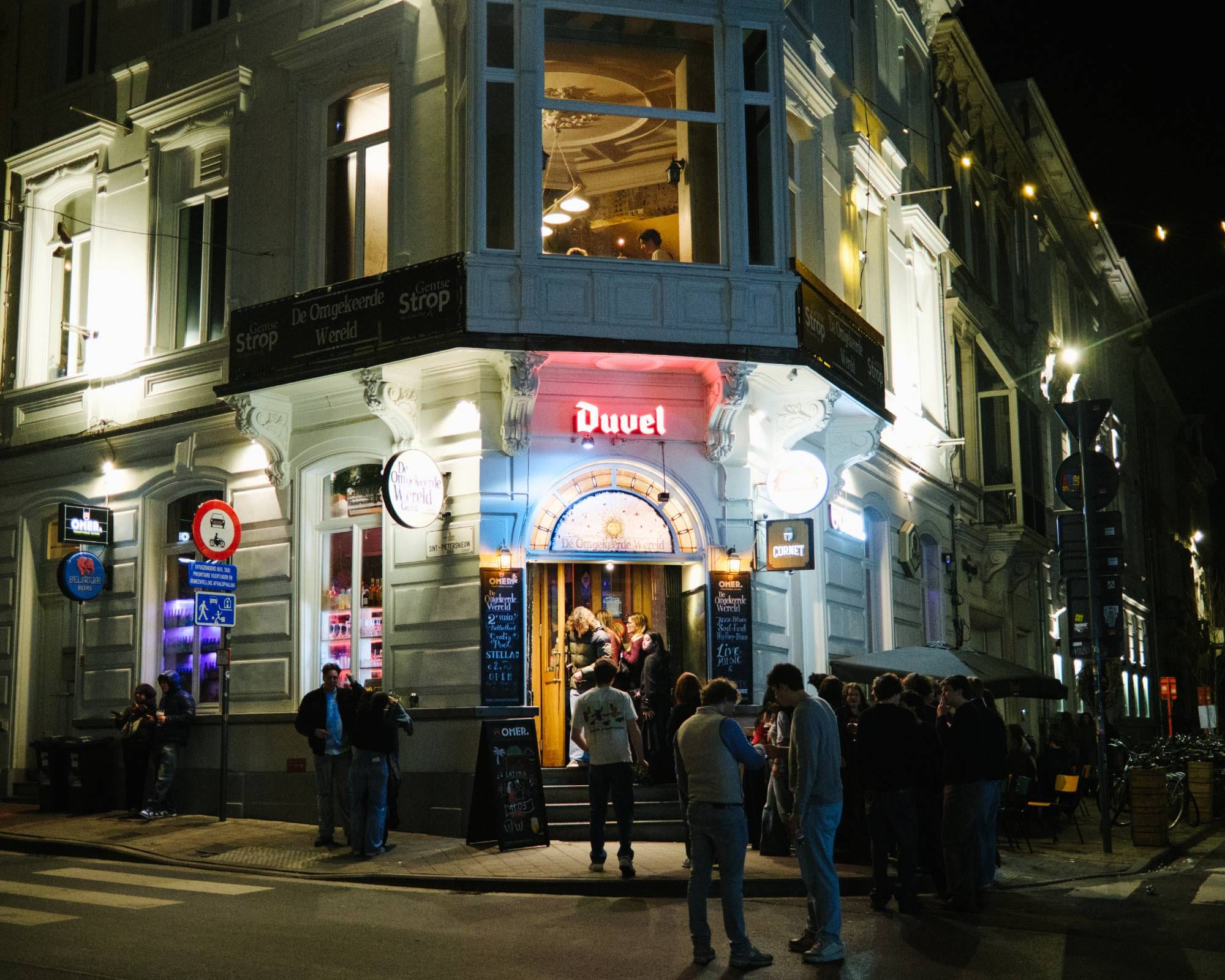 The corner facade of De Omgekeerde Wereld at night, with the red Duvel sign above the door and a crowd outside.