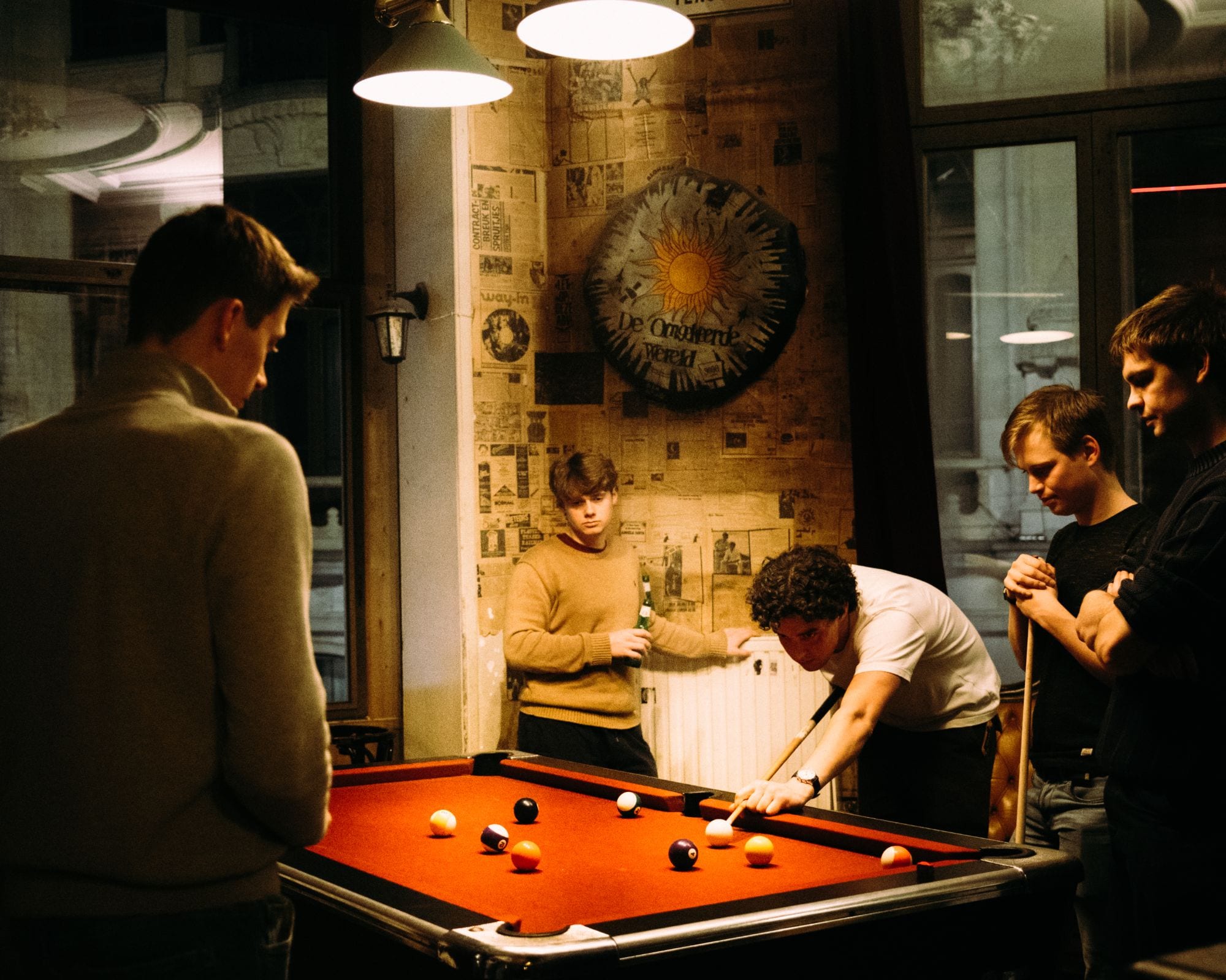 Friends around the red pool table under the lamps, with the sun of De Omgekeerde Wereld on the wall behind them.