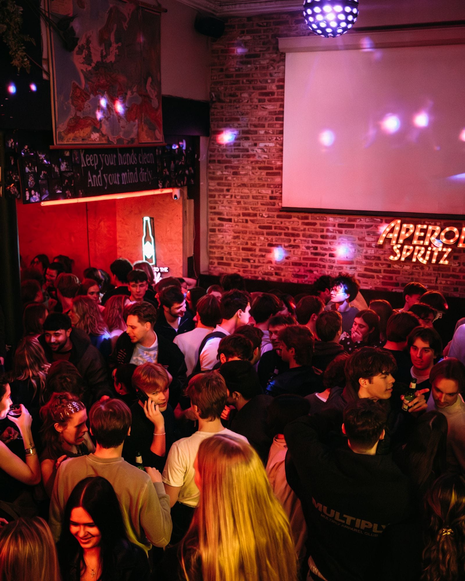 A packed room under a disco ball, with an Aperol Spritz sign on the brick wall.